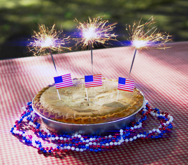 A Fourth of July apple pie with red, white, and blue beads, American flags, and sparklers on a picnic table with lens flare.